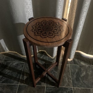 May include: A small wooden side table with a round top featuring a black mandala design. The table has a simple, cross-shaped base.