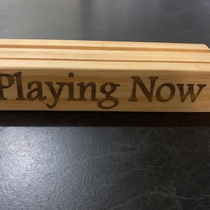 May include: A wooden sign with the text "Playing Now" engraved on it.