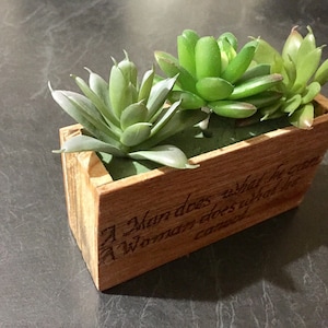 May include: A wooden planter box with a quote inscribed on the side: "A man does what he can. A woman does what a man cannot." The planter box contains three green succulent plants.