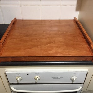 May include: A wooden countertop cover with raised edges and a light brown wood grain finish. The cover is placed on top of a white oven with a black countertop.