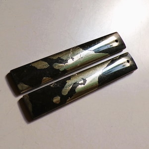 Op de afbeelding: Twee rechthoekige, gepolijste pyrietstenen met een donkere, metallic glans en onregelmatige goudkleurige insluitsels. Elke steen heeft een klein gaatje aan één uiteinde. De stenen liggen op een lichtgekleurd oppervlak.