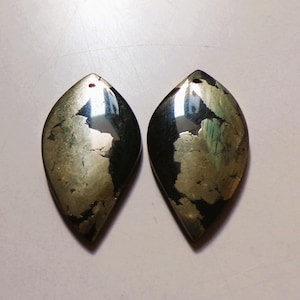 May include: Two pyrite gemstone pendants. Each pendant is a leaf shape with a hole at the top for stringing. The pyrite has a metallic luster with a mix of gold and black colors. The surface has a textured, cracked appearance.