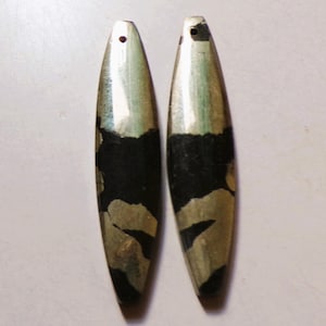 Puede incluir: Dos colgantes alargados con forma de lágrima. Tienen un brillo plateado-dorado con manchas negras irregulares. Cada colgante tiene un pequeño agujero en la parte superior, lo que sugiere que están diseñados para ser ensartados.