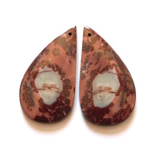 May include: Two teardrop-shaped stone pendants. The pendants have a reddish-brown base color with mottled patterns of dark red, green, and white. Each pendant has a small hole at the top for stringing.