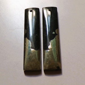 May include: Two rectangular, dark green pendants with a metallic, silver-colored bottom half. Each pendant has a small hole at the top. The pendants are displayed on a light-colored surface.