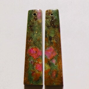 Könnte beinhalten: Zwei rechteckige Ohrringe mit floralem Muster. Die Ohrringe haben oben ein Loch zum Aufhängen. Das Design zeigt rosa Blüten, grüne Blätter und einen braunen Hintergrund. Die Ohrringe sind etwa 3,8 cm lang.