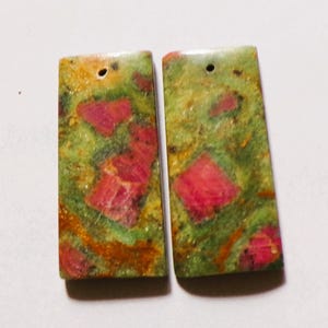 May include: Two rectangular unakite gemstone pendants. The pendants have a hole at the top for stringing. The unakite has a mottled appearance with green, pink, and orange hues. The pendants are ideal for jewelry making.