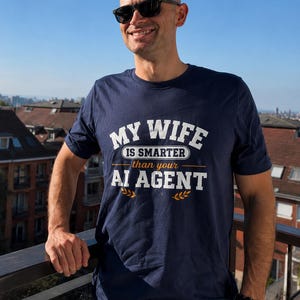 以下が含まれることがあります： ネイビーブルーの半袖Tシャツ。白とオレンジの文字で「MY WIFE IS SMARTER than your AI AGENT」とプリントされ、テキストの下には麦のような装飾アイコンが描かれています。ダークサングラスと腕時計を身につけた人物が着用しています。