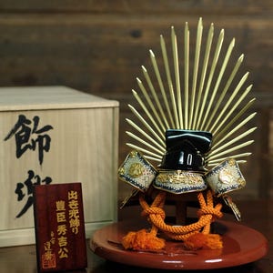 Puede incluir: Un casco de samurái japonés decorativo, con un cuenco lacado en negro, detalles dorados y una cresta en forma de abanico. El casco descansa sobre un soporte lacado en rojo con detalles de cuerda naranja. También hay una caja de madera y una pequeña placa de madera con texto japonés.