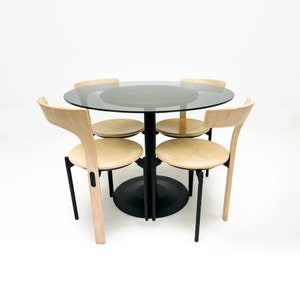 Puede incluir: Una mesa de vidrio negro con una base de metal negro y cuatro sillas de madera clara. Las sillas tienen un diseño sencillo con un respaldo curvo y un asiento redondo.