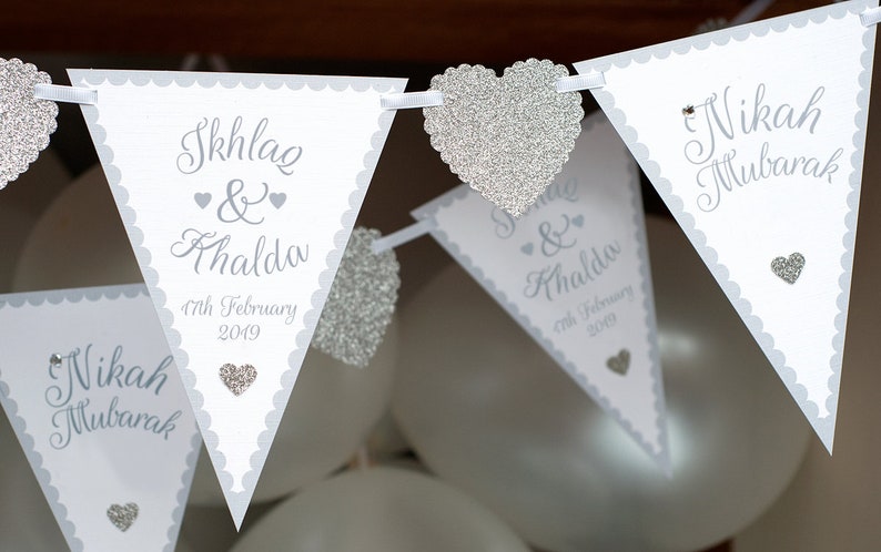 May include: A white and silver wedding banner with the text "Nikah Mubarak" and "Skhlag & Khalda" with the date "17th February 2019". The banner is decorated with silver glitter hearts.