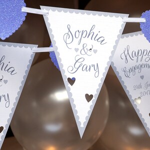 May include: White triangle pennant banner with gray scalloped edges and purple glitter hearts. The banner reads "Sophia & Gary" and "Happy Engagement 24th June 2017".