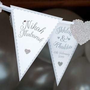 May include: White triangular pennant flags with scalloped edges and silver glitter hearts. The flags read "Nikah Mubarak" and "Ikhla & Ahalda 17th February 2019".