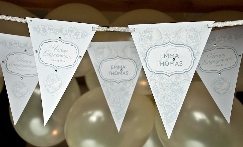 May include: A white and silver engagement party banner with a floral design. The banner has four flags with the text "Happy Engagement" and the date "12th June 2016" on two flags and the text "Emma & Thomas" on the other two flags.