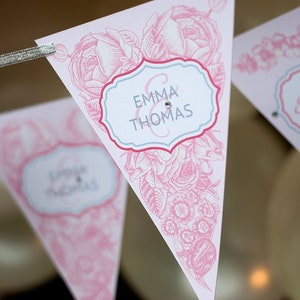 May include: Pink and white triangular pennant flags with floral designs and the text "Emma & Thomas" and "Happy Engagement".