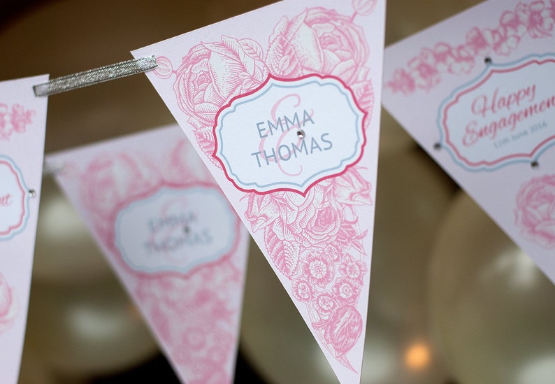 May include: Pink and white triangular pennant flags with floral designs and the text "Emma & Thomas" and "Happy Engagement".