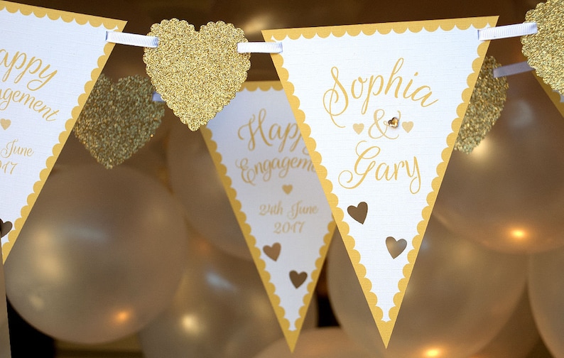 May include: White and gold engagement party banner with the text "Sophia & Gary" and "Happy Engagement 24th June 2017". The banner is decorated with gold glitter hearts.