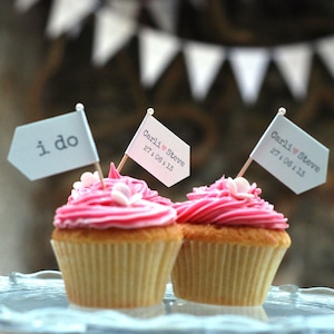 May include: Two pink frosted cupcakes with white sprinkles, decorated with flags. One flag says "i do" and the other says "Carli ♥ Steve 27.08.13".