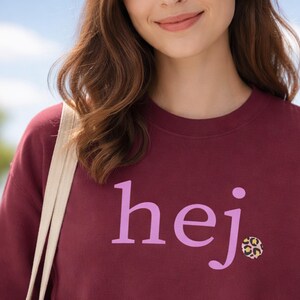 Puede incluir: Sudadera burdeos con la palabra "hej" en letras lilas. La letra "j" tiene un pequeño diseño de estampado de leopardo. La sudadera es de estilo cuello redondo.