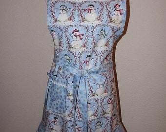 May include: A light blue apron with a repeating pattern of snowmen wearing hats and scarves. The apron has a ruffled hem and a tie at the waist. The background is a light blue with snowflakes.