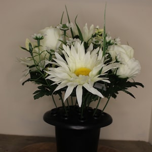 May include: A display of white flowers, including daisies and roses, with green leaves, arranged in a black vase. The central daisy has a yellow centre. The arrangement is a floral display.