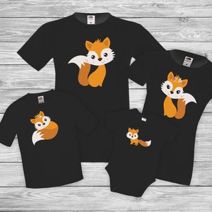 Könnte beinhalten: Ein Set aus vier schwarzen T-Shirts mit einem niedlichen Fuchs-Design. Die Shirts zeigen einen Fuchs mit orangefarbenem Fell und weißem Gesicht, der eine Blumenkrone trägt. Die Shirts sind perfekt für ein Familien-Matching-Outfit.