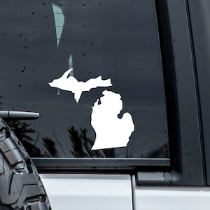 May include: White vinyl decal of the state of Michigan outline on the back window of a gray car. The decal is on the driver's side of the window.