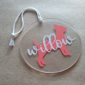 May include: A round, clear acrylic ornament with a pink silhouette of a dog and the name "Willow" written in white.