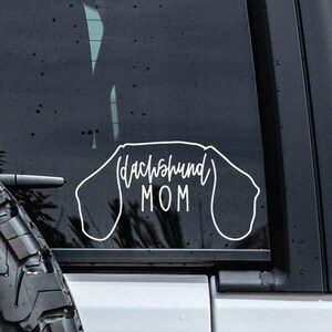 May include: A white vinyl decal with the text "Dachshund Mom" inside the outline of a dachshund dog. The decal is on the back window of a black car.