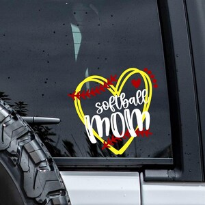 May include: A yellow heart-shaped decal applied to a vehicle window, featuring red baseball-style stitching accents and the white text "softball mom" written in a combination of cursive and bold sans-serif typography centered within the heart.