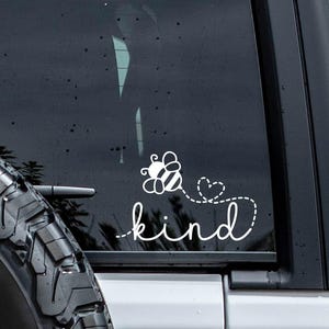 May include: White vinyl decal featuring a bee with a dotted trail leading to a heart, and the word "kind" in a cursive font. The decal is applied to the rear window of a black vehicle, with a spare tire visible.