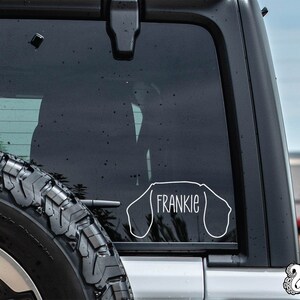 May include: A white vinyl decal of a dachshund dog with the name "Frankie" inside the outline. The decal is on the back window of a black car.
