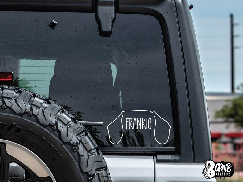 May include: A white vinyl decal of a dachshund dog with the name "Frankie" inside the outline. The decal is on the back window of a black car.
