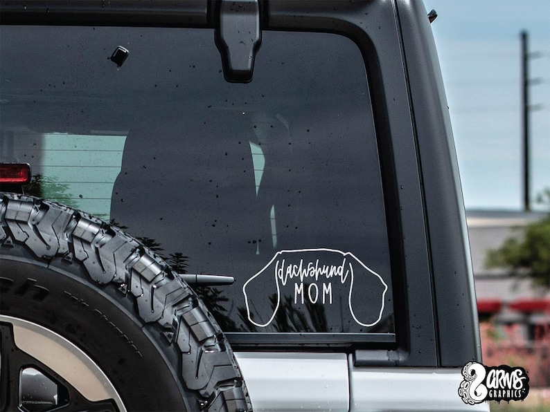 May include: A white vinyl decal with the text "Dachshund Mom" inside the outline of a dachshund dog. The decal is on the back window of a black car.