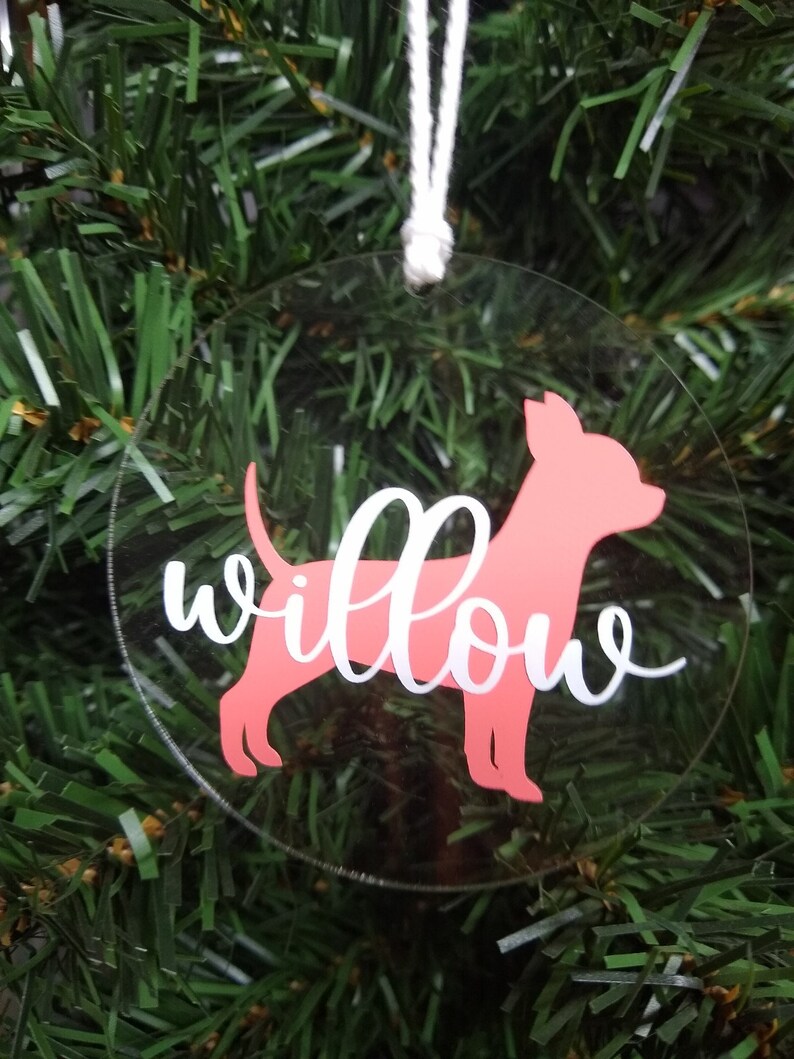 May include: A clear round Christmas ornament with a pink silhouette of a dog and the name "Willow" written in white cursive.