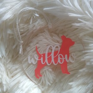 May include: A clear round ornament with a pink silhouette of a dog and the word "willow" written in white.