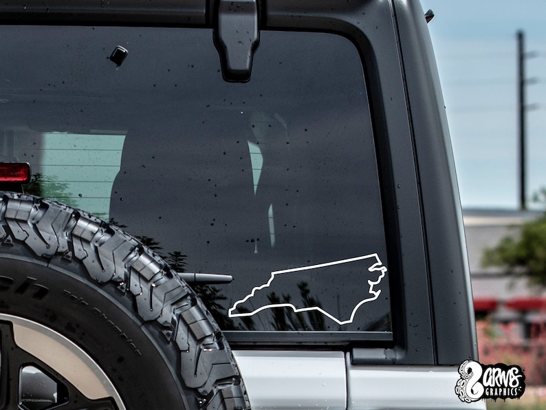 May include: A white outline of the state of North Carolina is displayed on the rear window of a black Jeep. The Jeep has a spare tire on the back.