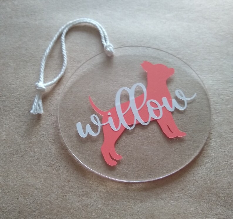 May include: A round, clear acrylic ornament with a pink silhouette of a dog and the name "Willow" written in white.