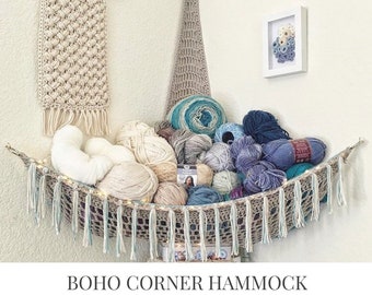 May include: A crocheted corner hammock with fringe, hanging in a corner with balls of yarn and a crocheted wall hanging. The text "BOHO CORNER HAMMOCK CROCHET PATTERN" is below the image.
