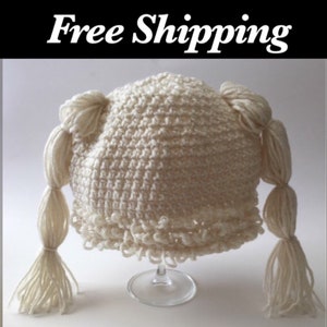 May include: White crocheted hat with two long yarn tails, resembling pigtails. The hat has a scalloped edge and a textured stitch pattern.