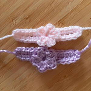 May include: Two handmade crochet bands, one in light pink and one in light purple, each featuring a central five-petaled flower motif. The bands are constructed with a simple stitch pattern and include extended yarn ends for tying or securing.
