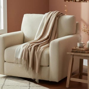 May include: A cream-colored armchair with a fringed beige and cream throw blanket draped over it. The chair is next to a small wooden side table with a mug, glasses, and books. The room has a neutral color palette.