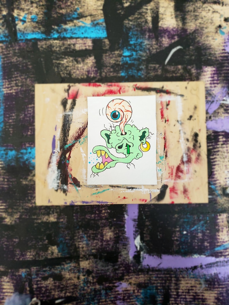 May include: A small, hand-drawn illustration of a green monster with a large, detached eyeball. The monster has yellow teeth, a gold earring, and blue and yellow accents. The artwork is on a white background, set against a textured, multi-coloured surface.