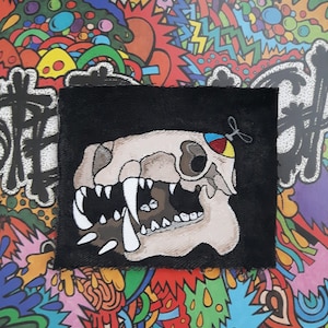 May include: A black fabric patch featuring a white and brown skull design. The skull has a large open mouth with sharp teeth and a colourful eye patch.