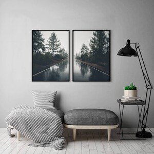 May include: Two black framed photographs of a foggy road through a forest. The road is wet and reflects the trees and sky. The photos are hung on a gray wall above a gray couch with a white and gray throw blanket.