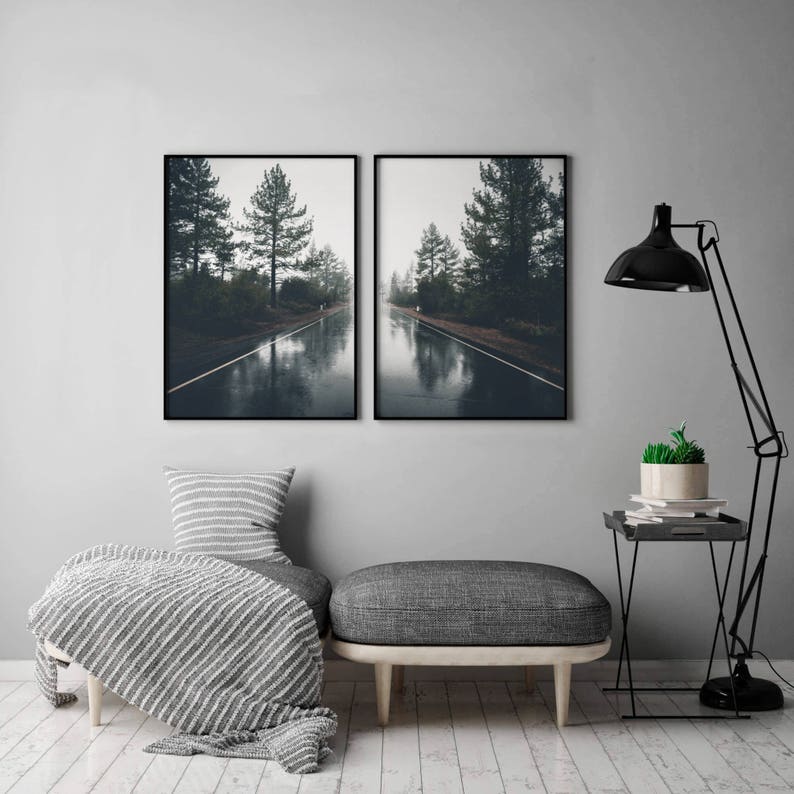 May include: Two black framed photographs of a foggy road through a forest. The road is wet and reflects the trees and sky. The photos are hung on a gray wall above a gray couch with a white and gray throw blanket.