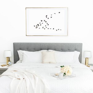 K&ouml;nnte beinhalten: Ein wei&szlig;es Bett mit einem grauen Kopfteil und wei&szlig;en Bettw&auml;sche. Eine wei&szlig;e Decke ist am Fu&szlig;ende des Bettes gefaltet. Ein wei&szlig;es Tablett mit einer wei&szlig;en Tasse, einem Teller mit Keksen und einem Strau&szlig; wei&szlig;er Blumen steht auf dem Bett. Ein gerahmter Druck eines Vogelschwarms im Flug h&auml;ngt &uuml;ber dem Kopfteil.