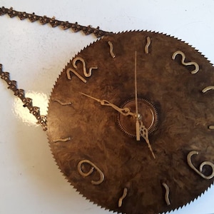 Puede incluir: Un reloj hecho de una hoja de sierra circular con números y manecillas doradas. El reloj está colgado de una cadena.