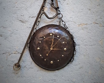 May include: A rustic metal clock with a round face and Roman numeral markings. The clock is suspended from a metal chain and hook, resembling an old-fashioned scale.