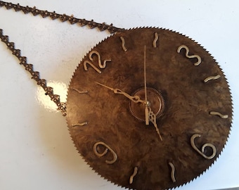May include: A clock made from a circular saw blade with gold-colored numbers and hands. The clock is hanging from a chain.